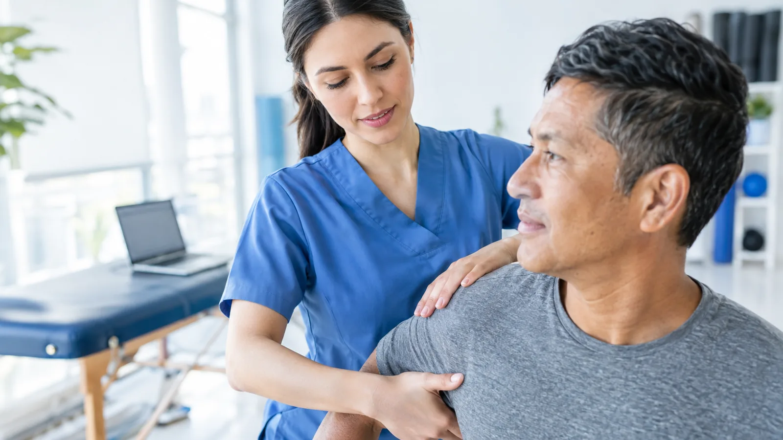 A physical therapist assesses a patient's shoulder with a laptop in the background